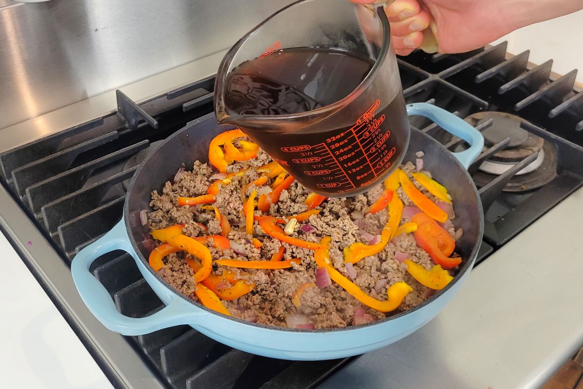 This is an image of a hand holding a container of beef broth, over a skillet of cooked ground beef and vegetables. 