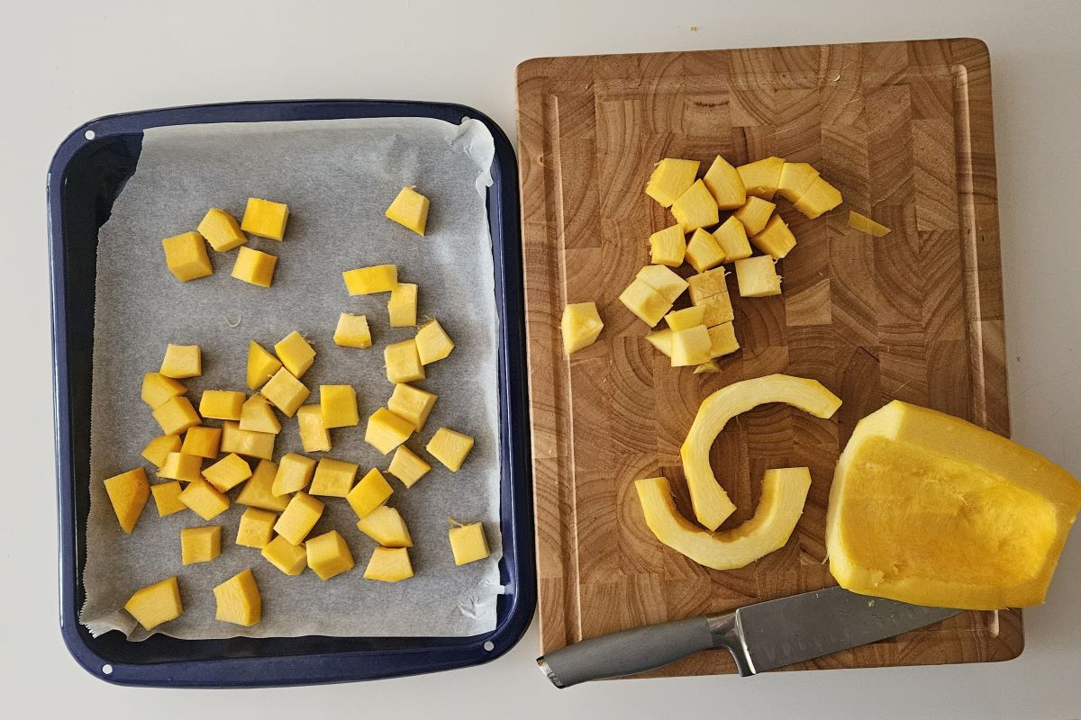 This is an image of a cutting board with cubed squash. Beside it, there is a roasting pan with pieces of squash. 
