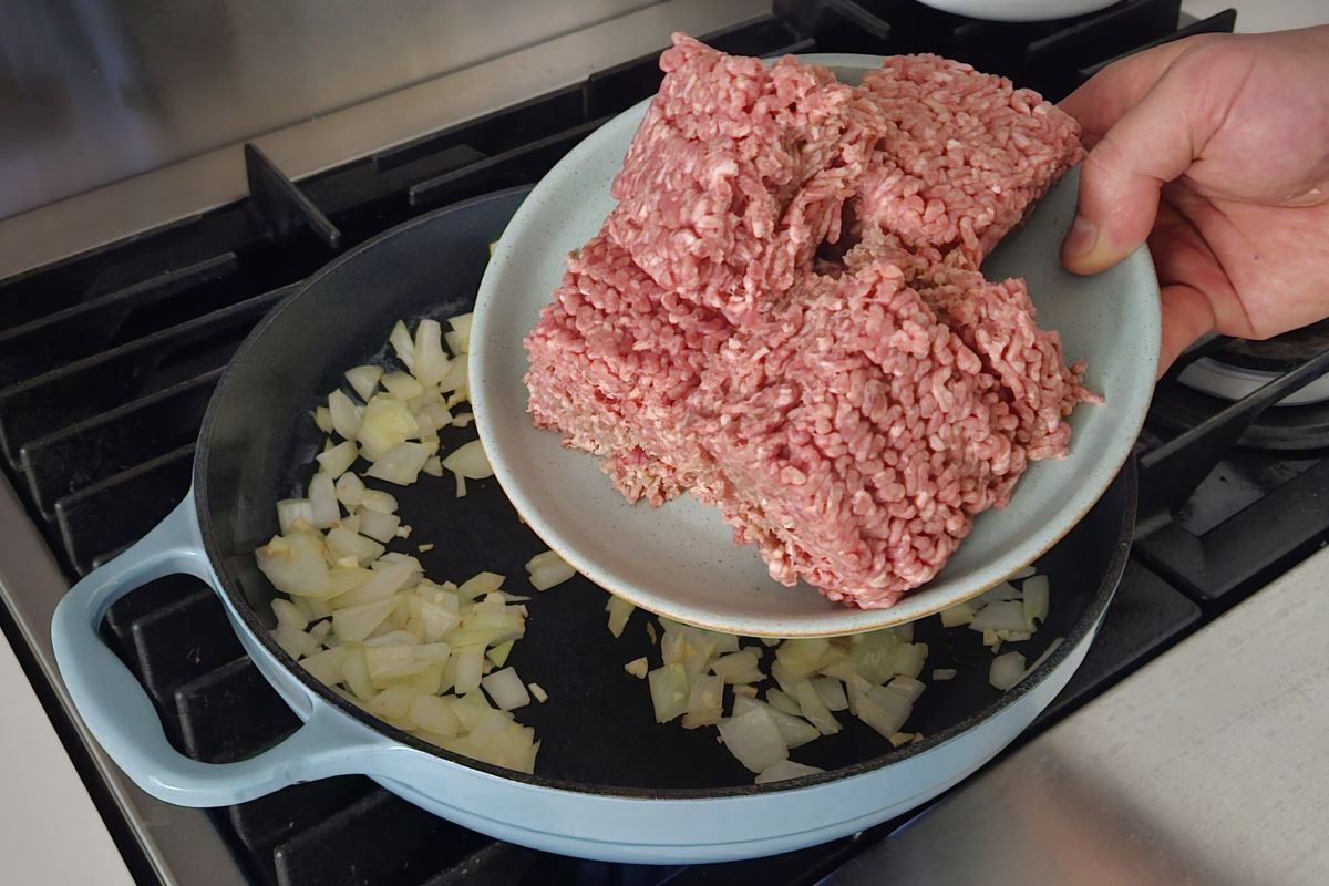 Someone placing ground meat in a large skillet with onions. 
