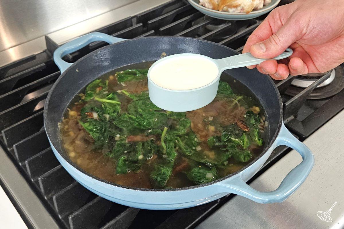 Someone holding a measuring cup of heavy cream over a skillet.