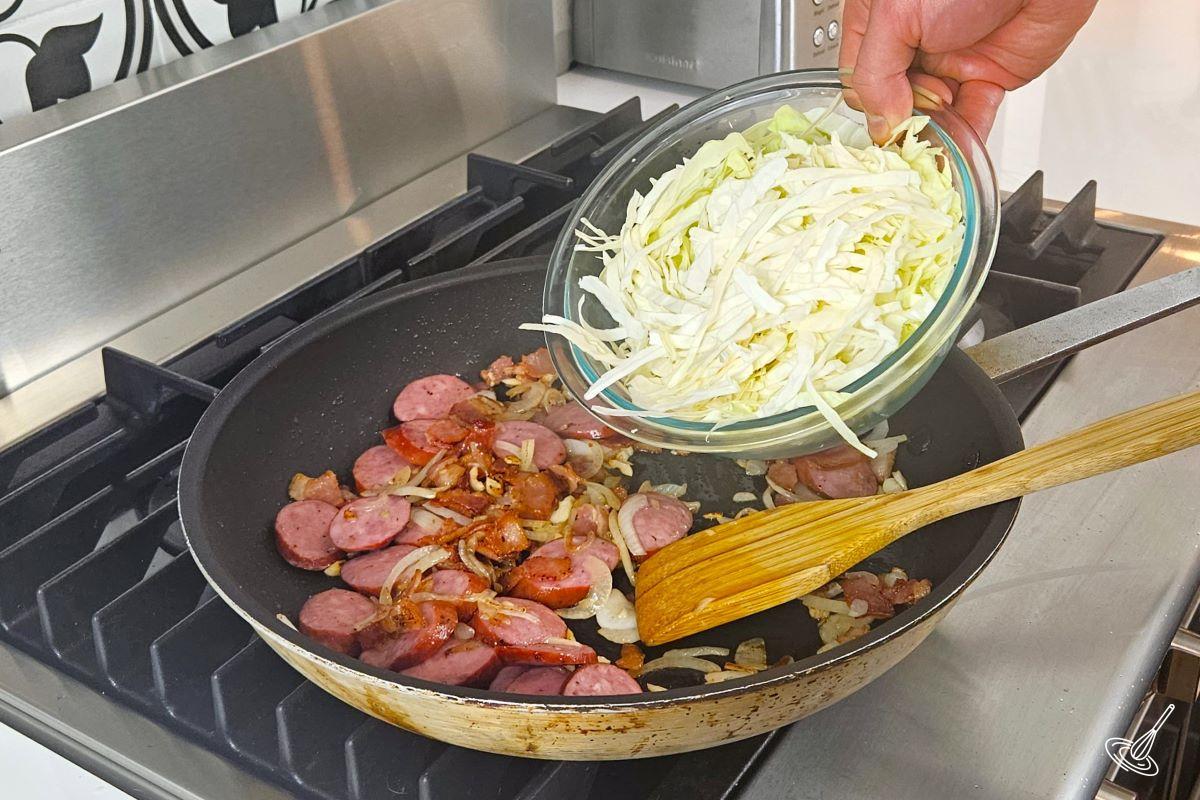 Someone placing shredded cabbage in a large frying pan, which contains farmers sausage, onions, garlic, and bacon.