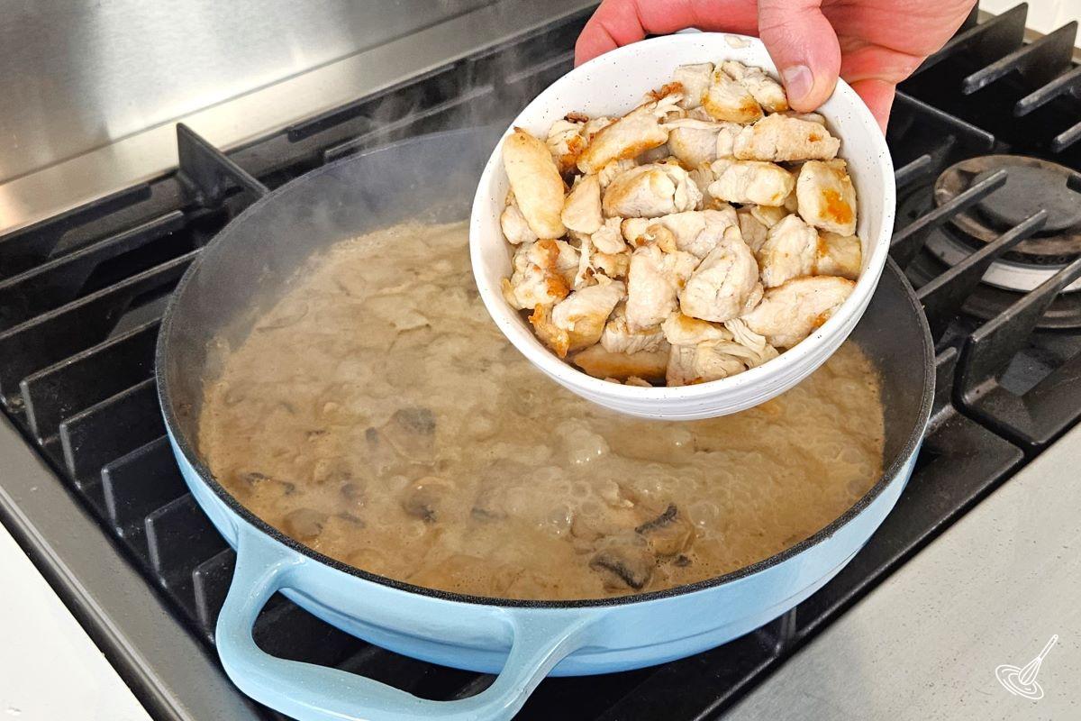 Someone placing a bowl of cooked chicken pieces in a large skillet with a brown sauce.
