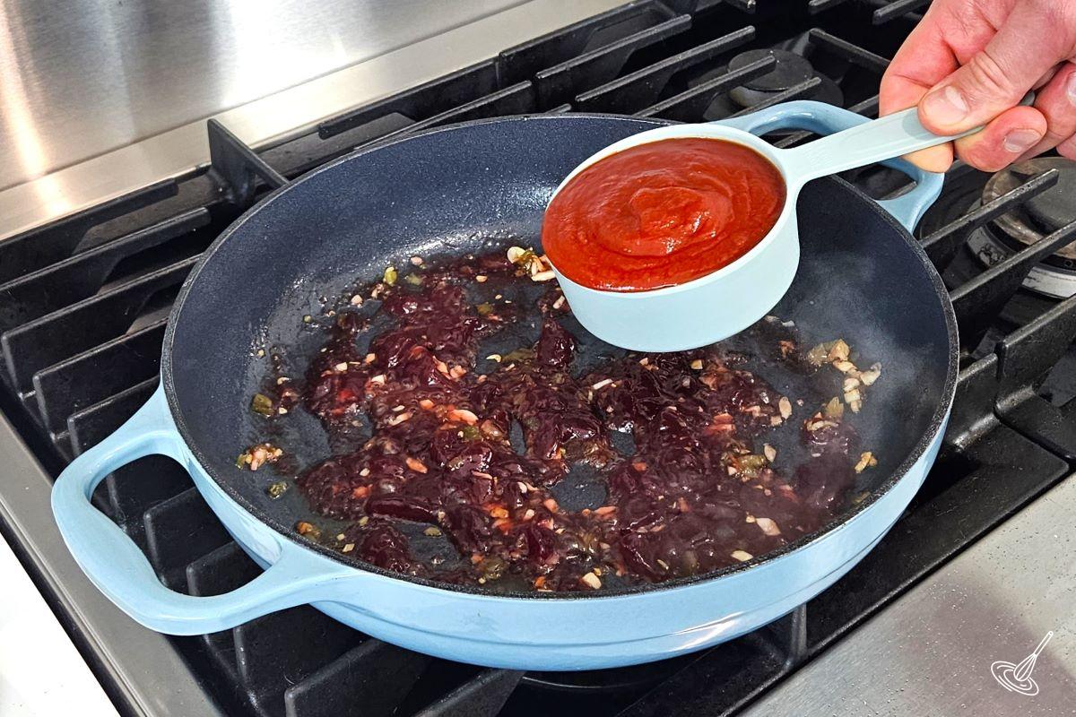 Someone placing chili sauce in a large skillet with cranberry sauce, garlic and jalapenos.