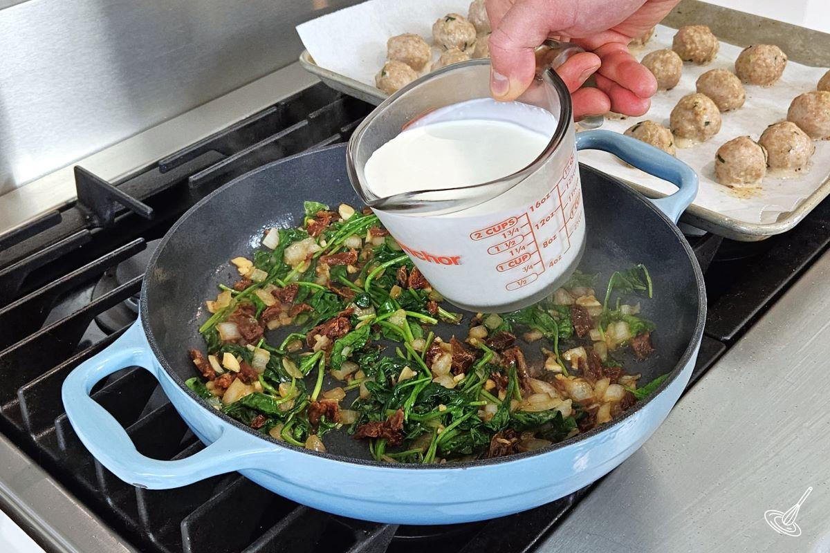Someone pouring heavy cream into a large skillet with broth and vegetables. 