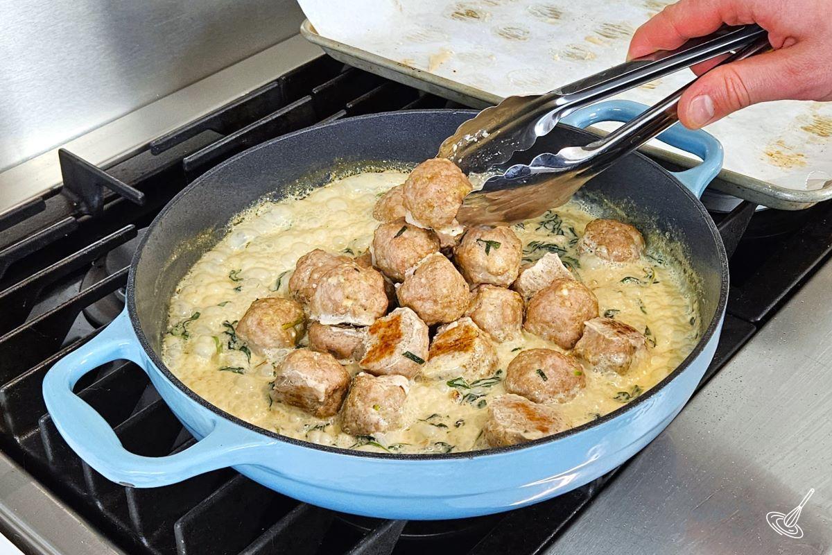 Someone placing cooked meatballs into a large skillet containing a creamy vegetable sauce. 