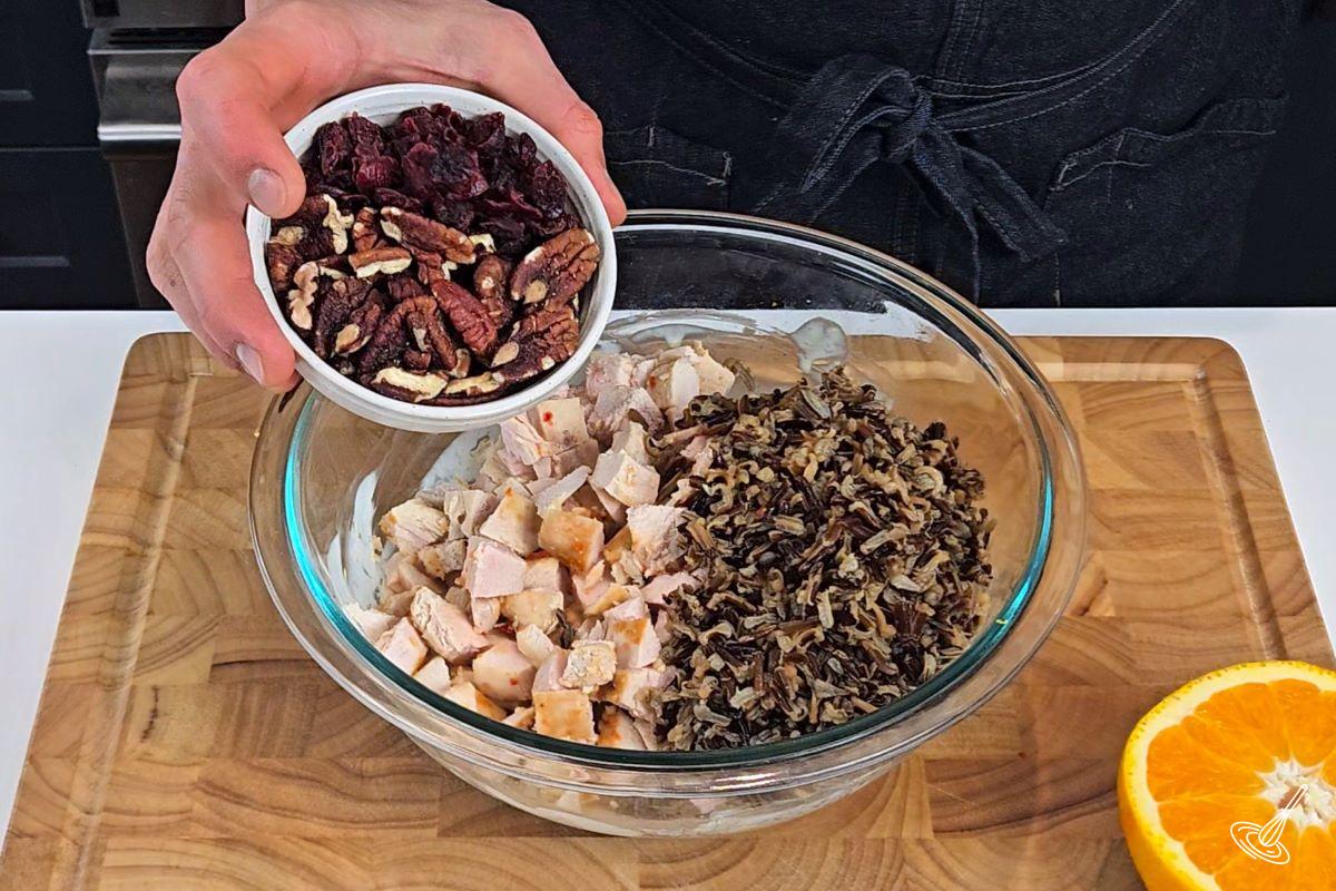 Someone placing craisins and pecans in a large bowl with cubes chicken and wild rice. 