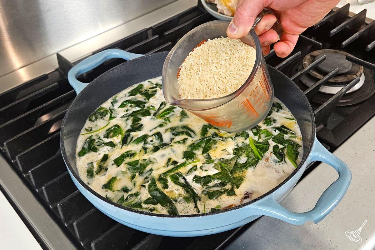 Someone pouring uncooked rice in a skillet which contains broth and vegetables.