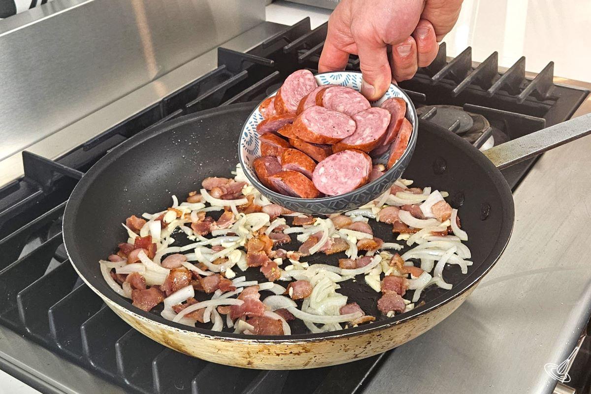 Someone placing sliced farmers sausage into a frying pan with onions, garlic, and bacon.