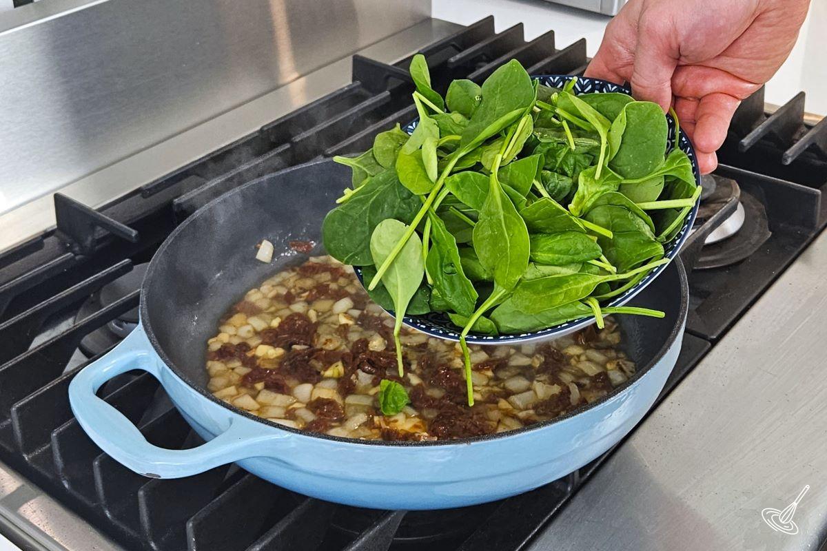 Someone placing fresh spinach in a large skillet with sundried tomatoes, onion, garlic, and broth. 