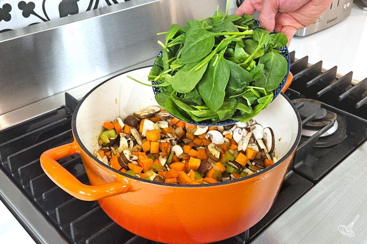 Someone adding fresh spinach into a Dutch oven containing other chopped vegetables. 