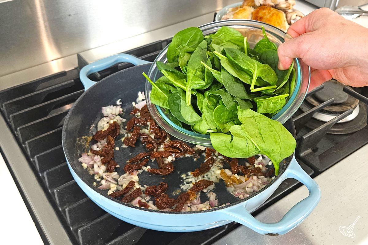 Someone placing fresh spinach leaves in a skillet to cook with shallots and garlic.