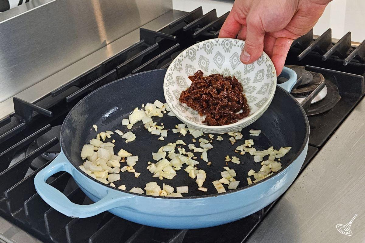 Someone placing sundried tomatoes in a large skillet with onions and garlic.