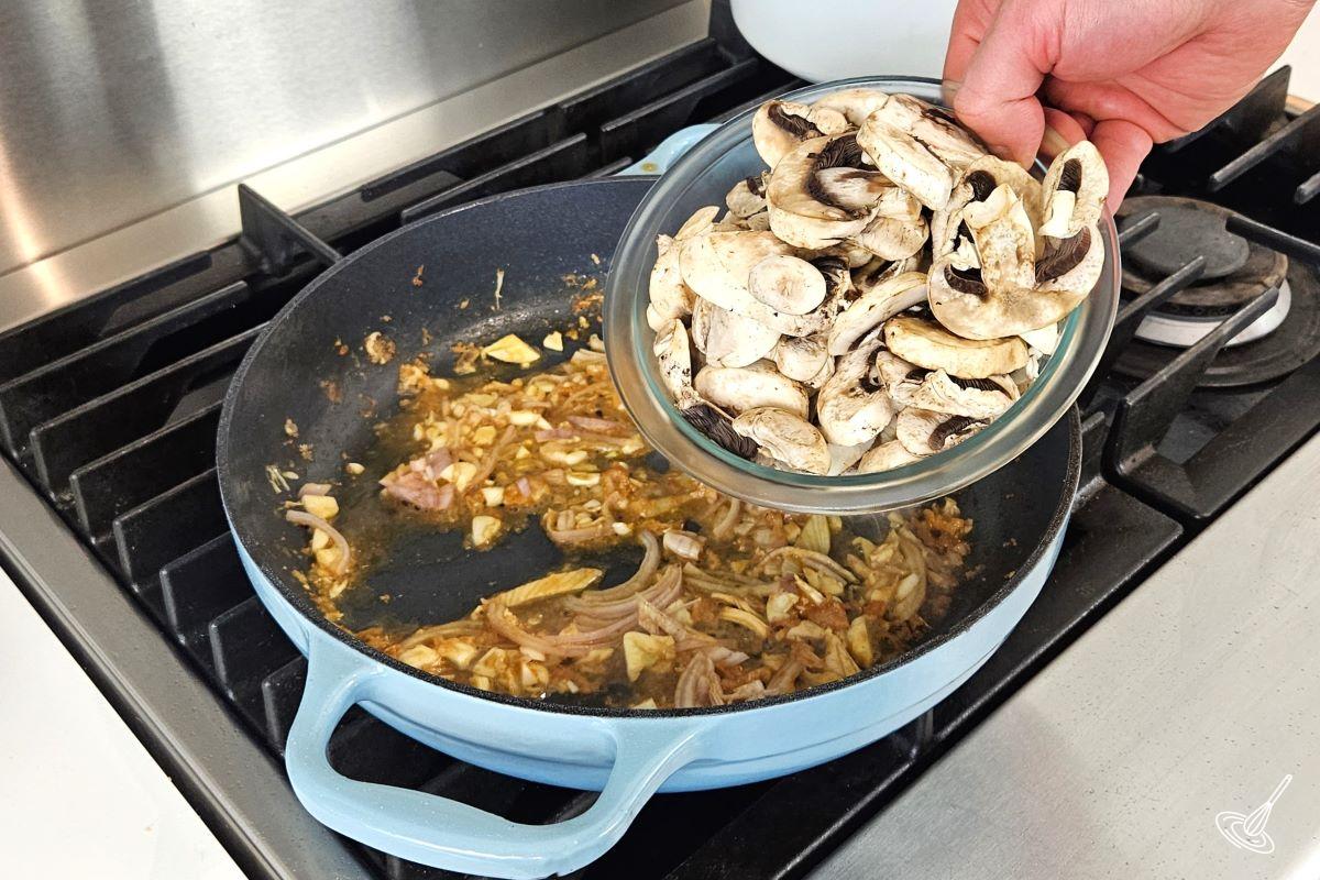 Someone placing sliced mushrooms in a large skillet with cooked shallots and garlic.