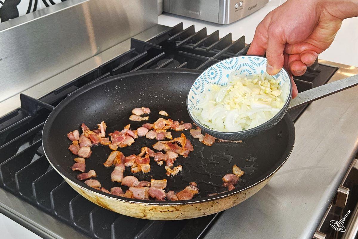 Someone placing chopped onion and garlic in a frying pan with chopped bacon.