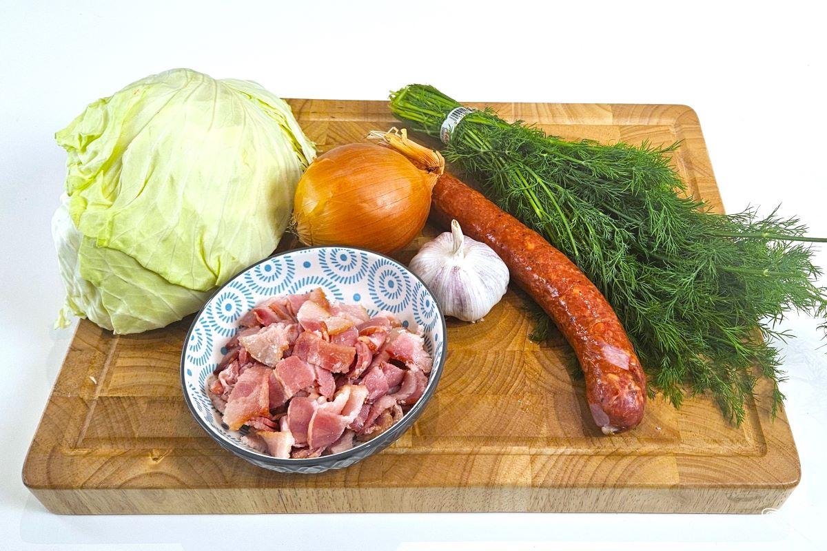 Chopped bacon, farmers sausage, garlic, onion, fresh dill and a cabbage head, on a wooden cutting board.