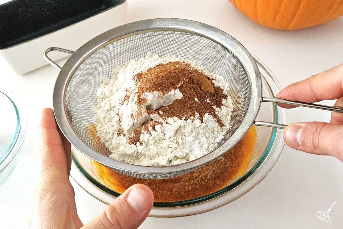 Someone sifting flour and other dry ingredients in a large bowl with pumpkin puree and other wet ingredients.