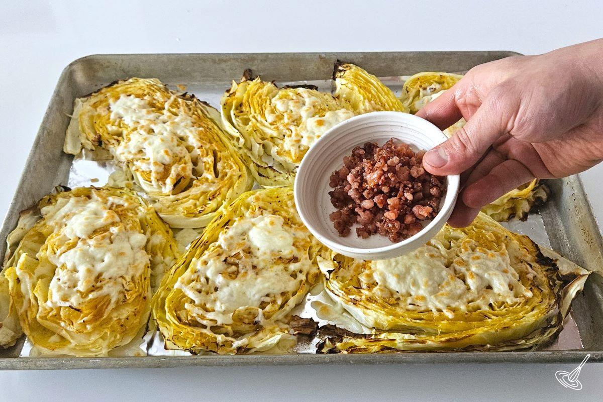Someone holding a bowl of bacon bits over a baking sheet of Cheesy Cabbage Steak.