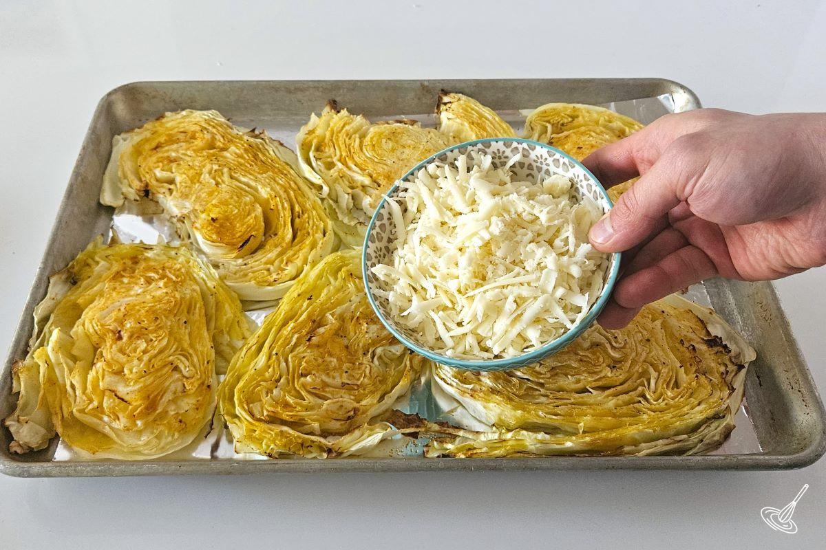 Someone holding a small bowl of grated cheese over a tray of roasted cabbage.