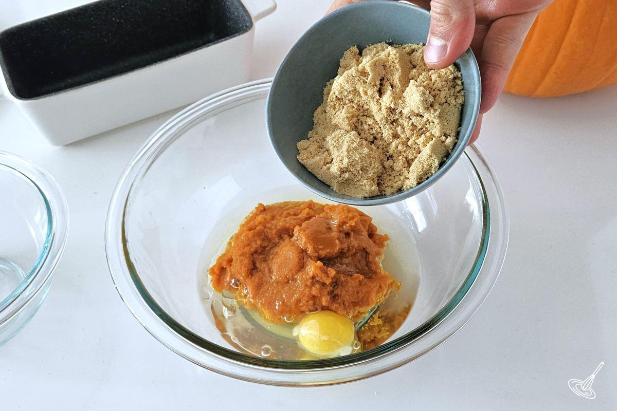 Someone placing brown sugar in a large bowl with pumpkin puree, egg and vanilla.