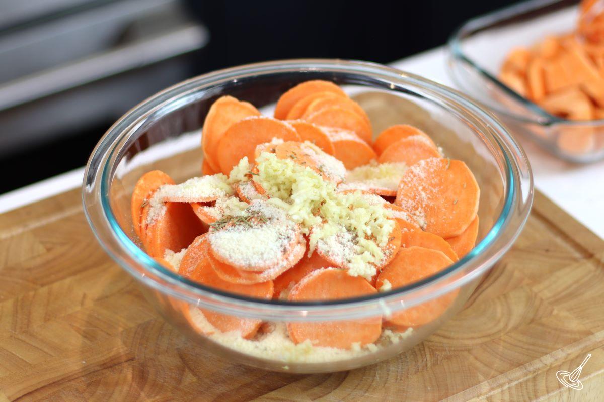 Slice of sweet potato in a large bowl with crushed garlic, rosemary, and parmesan cheese. 