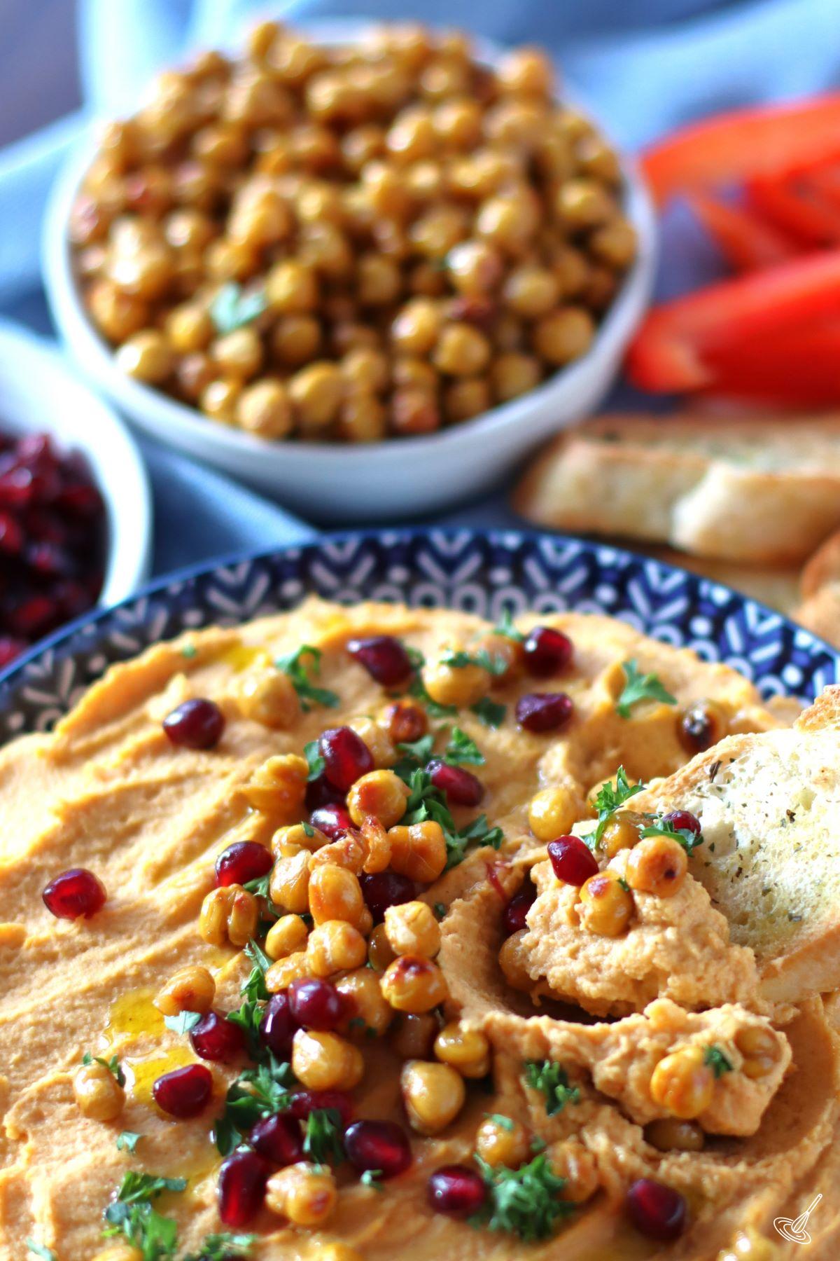 A bowl of hummus, topped with pomegranate seeds and Honey Roasted Chickpeas. 