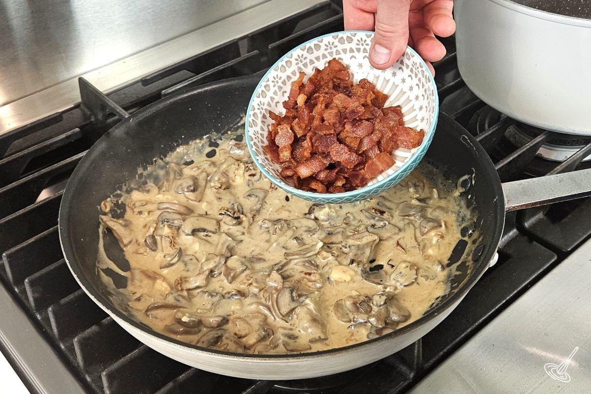 Someone adding bacon back into the frying pan with vegetables and sauce. 