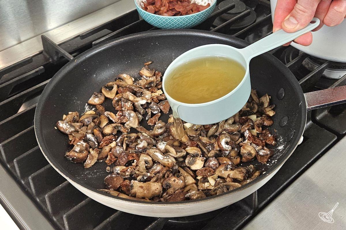 Someone pouring in a cup of chicken broth in a frying pan with vegetables. 