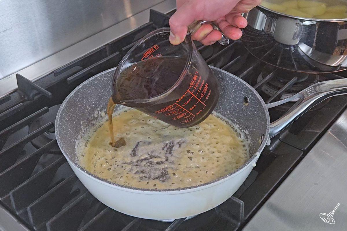 Someone adding beef broth to a large skillet with a roux. 