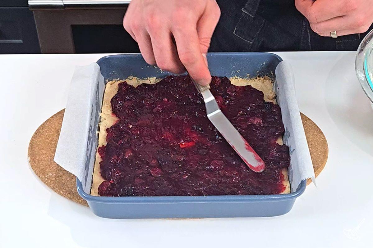 Someone spreading cranberry sauce in a baking pan, on top of dough. 