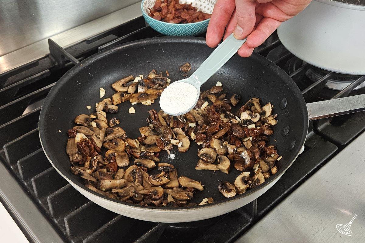 Someone sprinkling flour in a large frying containing vegetables.