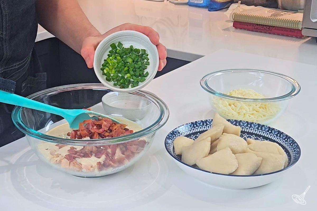 Someone placing chopped green onion into a bowl with bacon bits and ranch dressing.