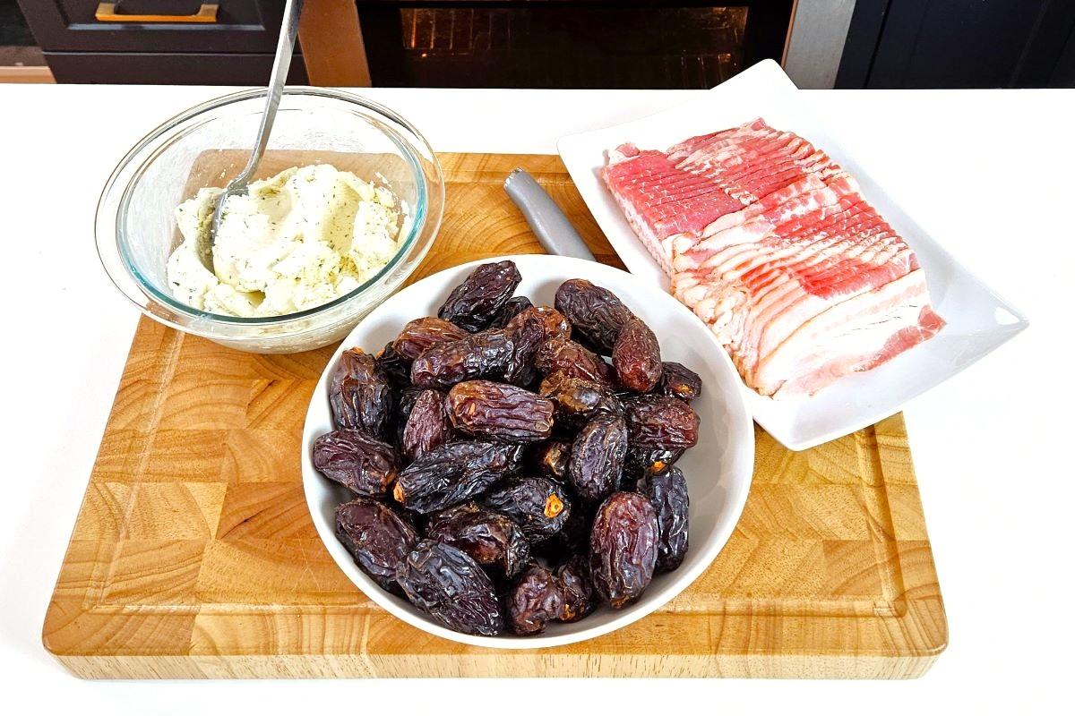 A bowl of Medjool dates, a plate of bacon slices, and goat cheese filling, on top of a cutting board.