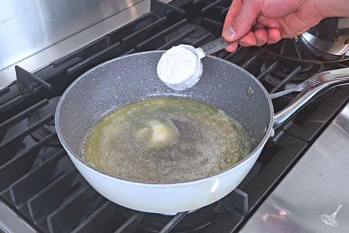Someone placing flour in a large skillet with melted butter.