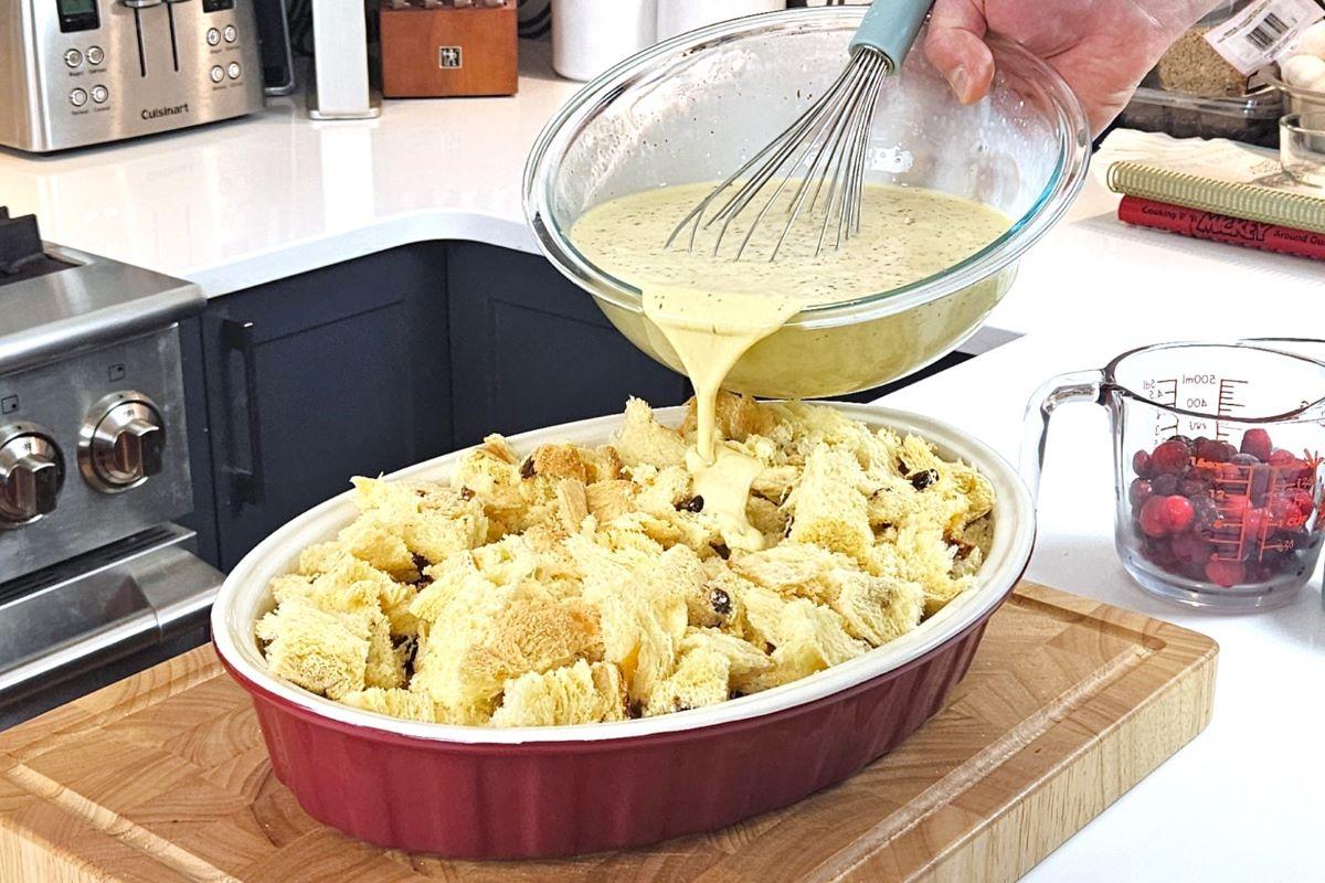Someone pouring an eggnog mixture into a baking dish containing cubed bread.