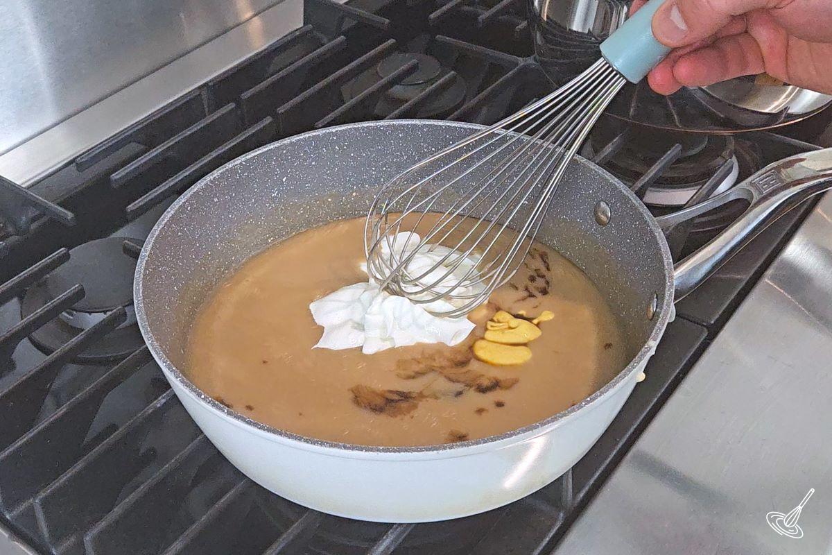Someone whisking sour cream into gravy in a large skillet. 