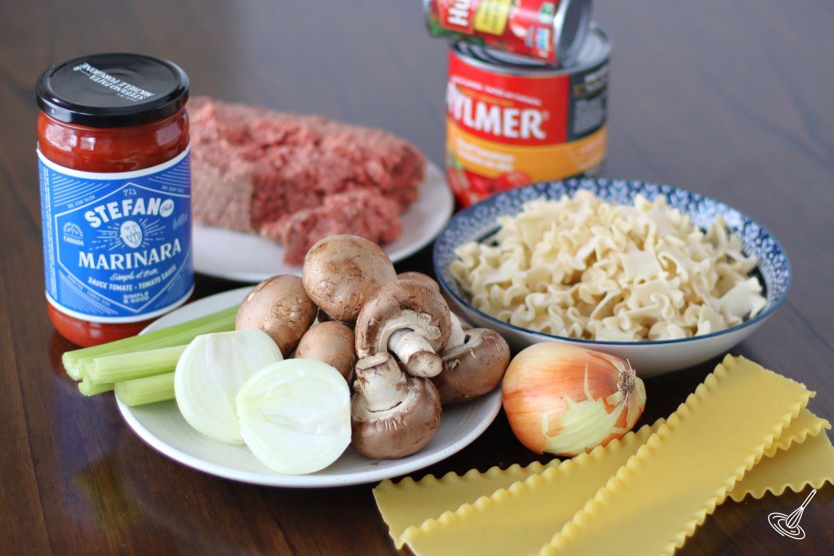 Ingredients laid out on a table, including fresh vegetables, uncooked lasagna pasta, ground beef, and tomato sauce.