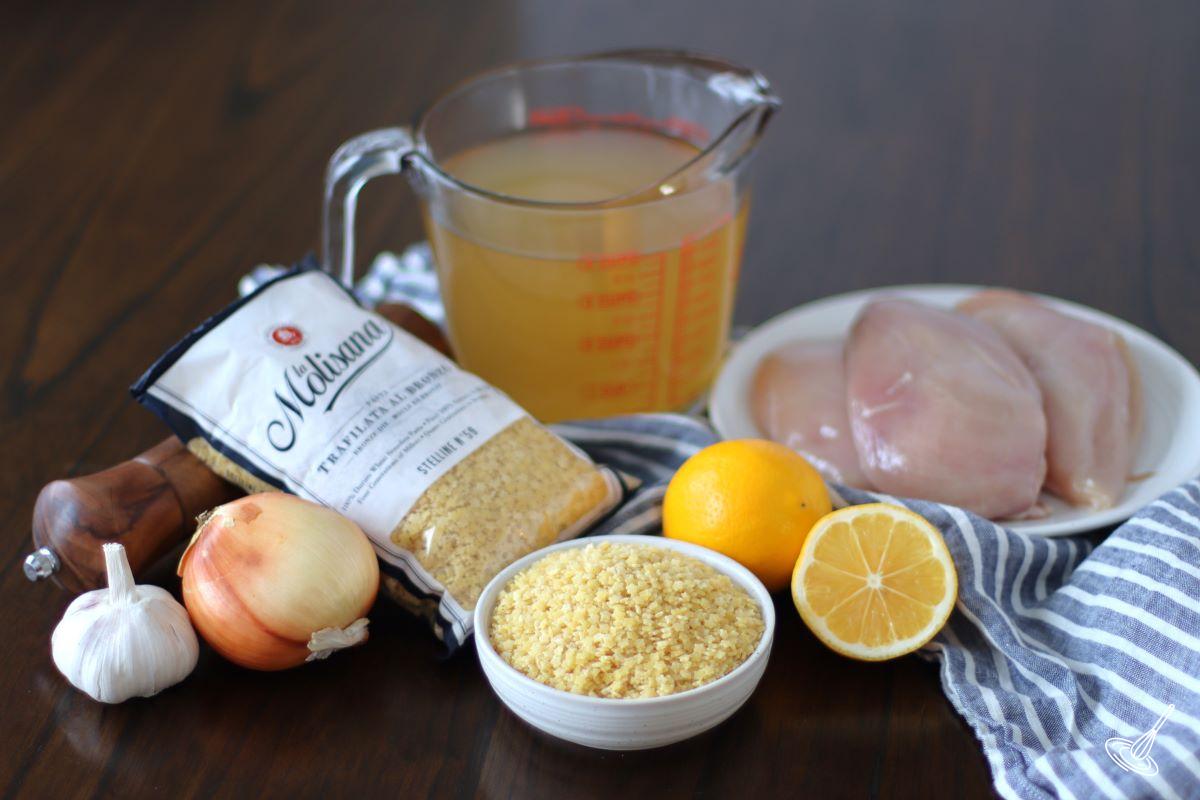 Ingredients laid out on a table, including chicken breasts, chicken broth, lemons, and pasta.