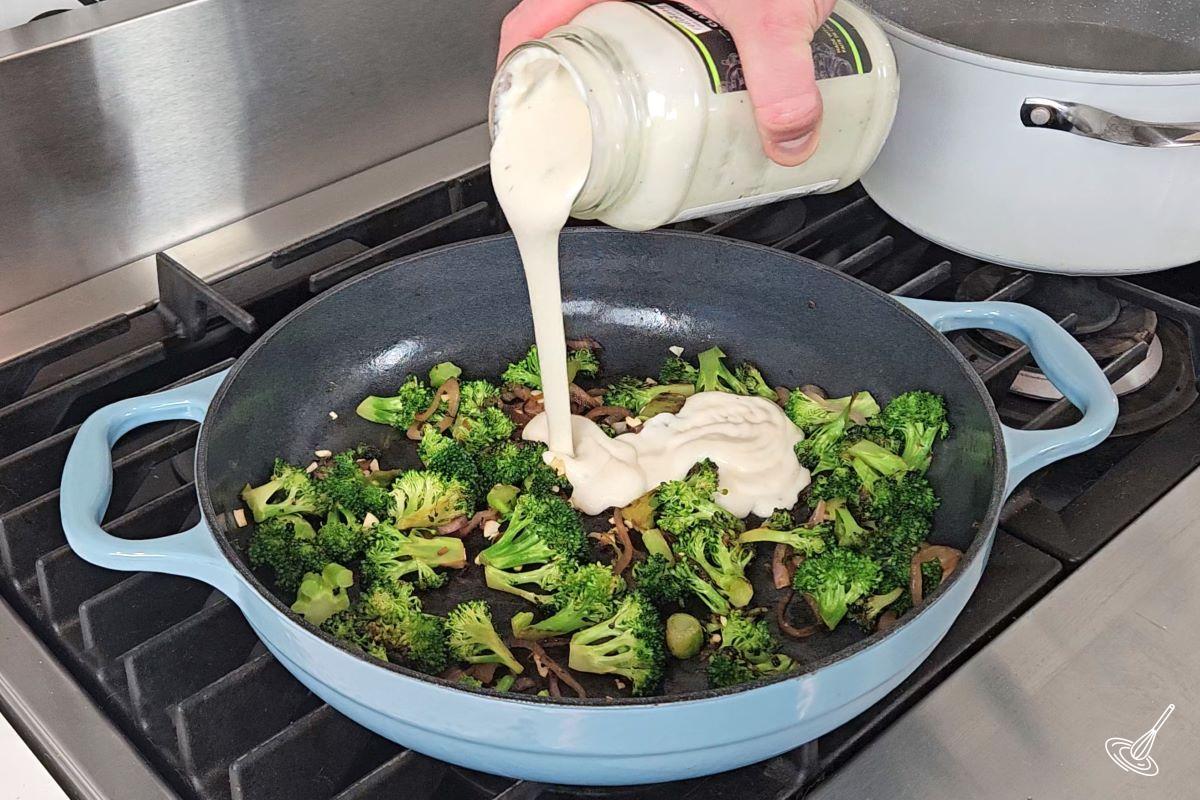 Someone pouring alfredo sauce in a large skillet containing cooked vegetables. 
