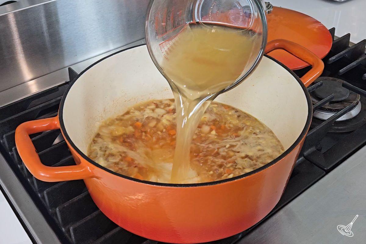 Someone deglazing a Dutch oven by slowly pouring in chicken broth. 