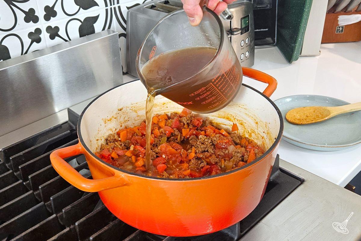 Someone pouring beef broth into a large pot of cooked ground beef and tomatoes. 