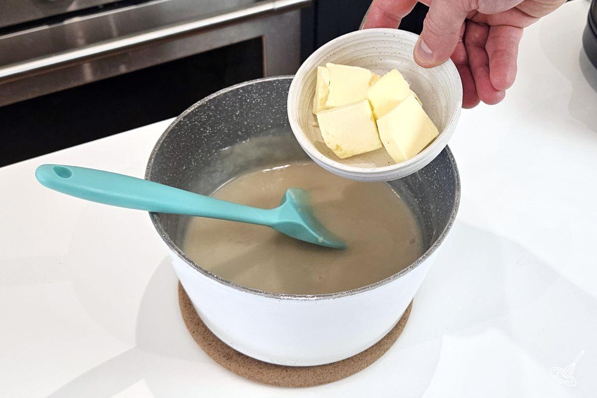 Someone placing cubed butter in a saucepan containing waffle sauce.