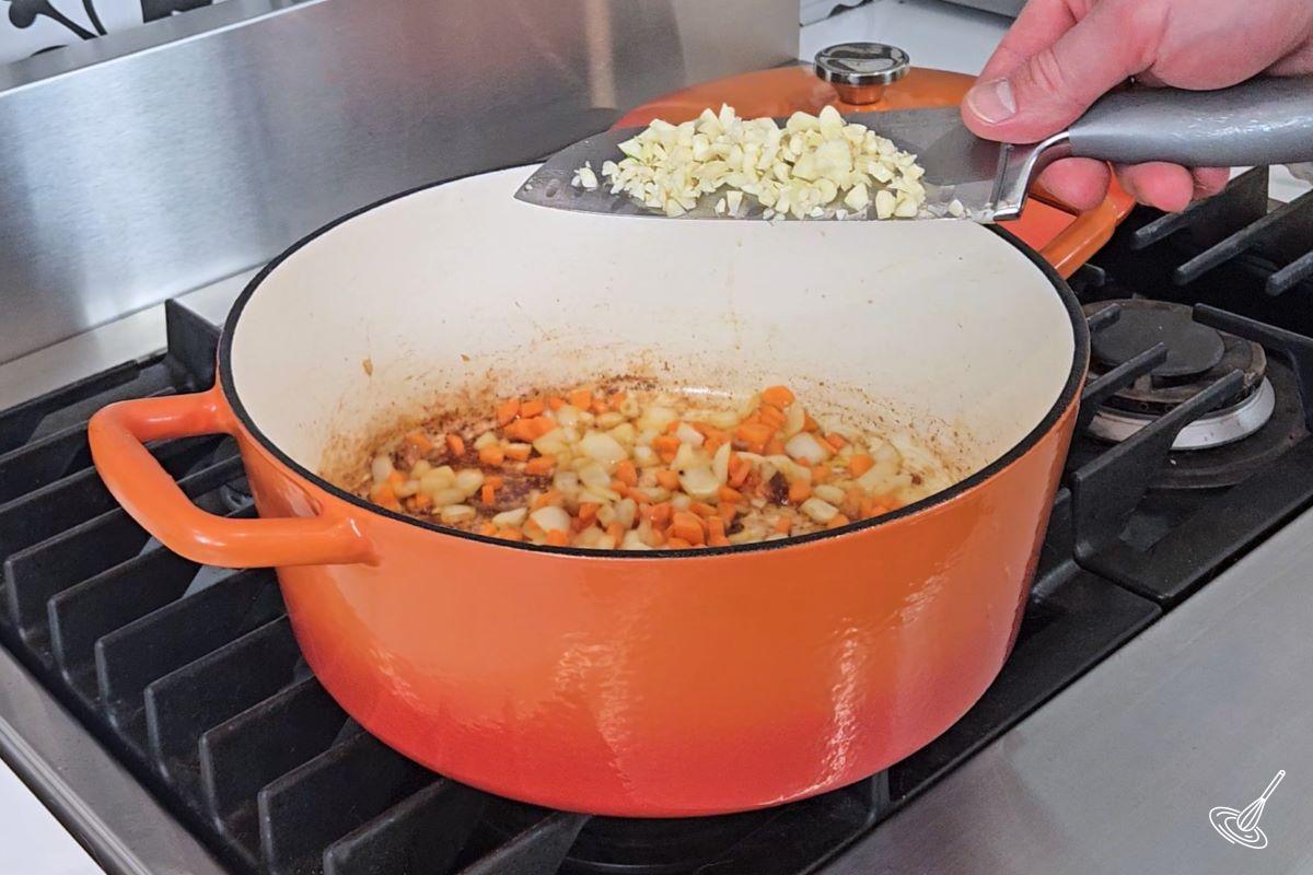 Someone adding chopped garlic into a Dutch oven with carrots and onions.