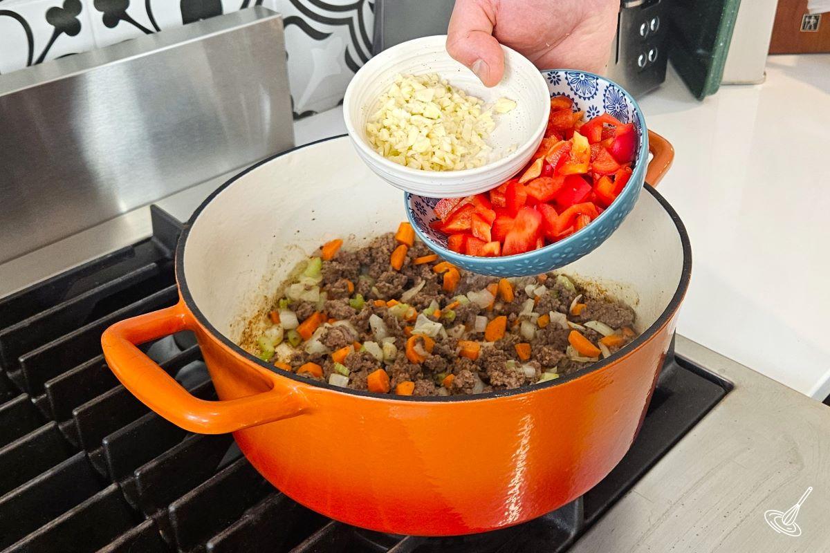 Someone placing garlic and bell peppers in a Dutch oven containing ground beef.