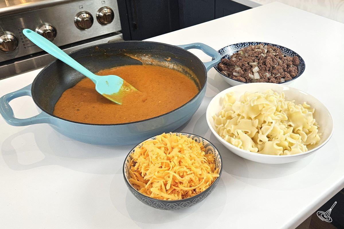 A large skillet of tomato sauce on the counter with separate bowls of grated cheese, cooked pasta, and cooked ground beef. 