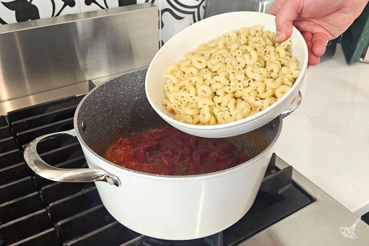 Someone adding cooked elbow macaroni to a large pot containing canned tomatoes and veggies.