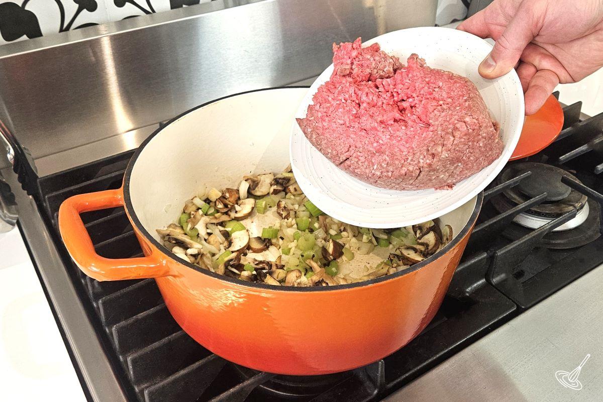 Someone placing ground beef in a Dutch oven with cooked vegetables.