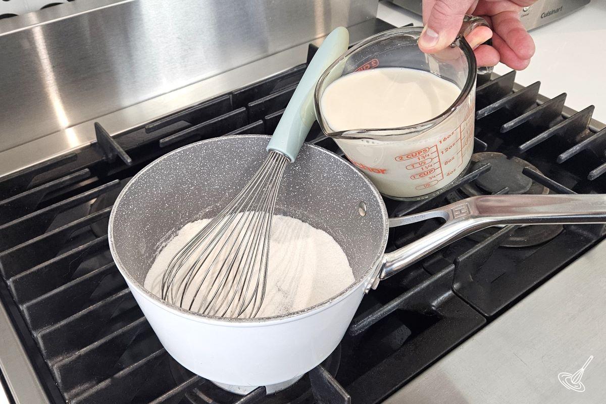Someone pouring milk in a saucepan which contains flour and sugar. 