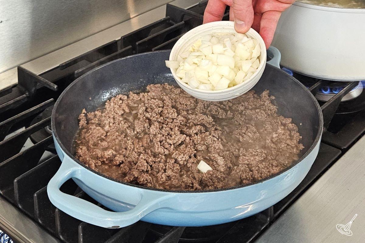 Someone placing onion and garlic in a large skillet with cooked ground beef.