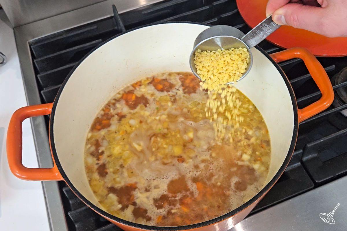Someone sprinkling small pasta into a Dutch oven which contains soup. 