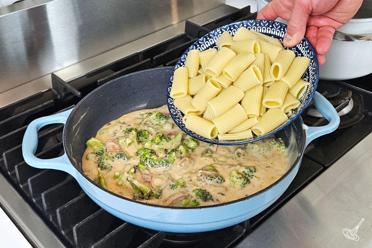 Someone adding cooked pasta to a large skillet containing Cajun Alfredo sauce with vegetables. 